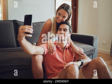 Heureux jeune couple caucasien assis ensemble et prenant selfie avec un smartphone. Jeune homme tenant un téléphone portable pour prendre des photos en étant assis entre les deux Banque D'Images