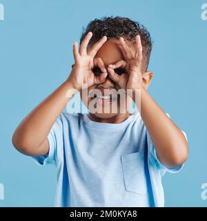 Portrait d'un petit garçon curieux qui regarde à travers les doigts en forme de jumelles contre un fond bleu de studio. Enfant curieux explorant Banque D'Images