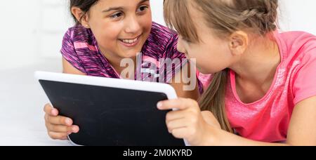 Deux petites filles mignonnes utilisent une tablette numérique et sourient lorsqu'elles sont allongées sur le lit dans la chambre des enfants Banque D'Images