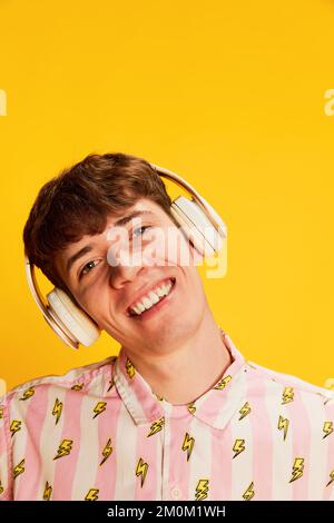 Portrait en gros plan d'un jeune homme émotif dans un casque souriant, en écoutant de la musique isolée sur fond jaune vif. Culture de la jeunesse, positive Banque D'Images