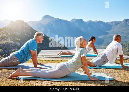 Groupe d'aînés divers pratiquant la posture du cobra pendant la formation de groupe au yoga. Personnes matures s'exerçant ensemble dans la nature avec vue sur la montagne dans Banque D'Images