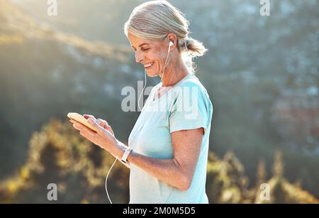 Femme sénior active qui porte des écouteurs et utilise une application mobile lors d'une séance d'entraînement dans la nature par temps ensoleillé Banque D'Images