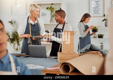 Deux concepteurs de vêtements utilisant un ordinateur portable tout en travaillant avec des matériaux au travail. Une jeune femme afro-américaine s'adapte à la conversation tout en travaillant avec une femme Banque D'Images