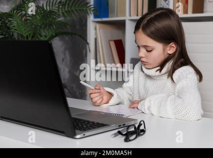 Grave enfant d'école de brunette caucasien étudiant à la maison. une fille est assise au bureau avec un ordinateur portable, écrire dans un ordinateur portable, faire ses devoirs ou se rendre Banque D'Images