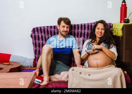 homme adulte caucasien barbu assis à côté de sa femme enceinte une femme brésilienne dans le salon de repos, ils sont souriants et regardent la caméra Banque D'Images