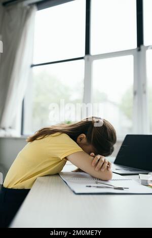 vue latérale d'une écolière épuisée qui dormait près d'un ordinateur portable avec écran vierge sur la table Banque D'Images