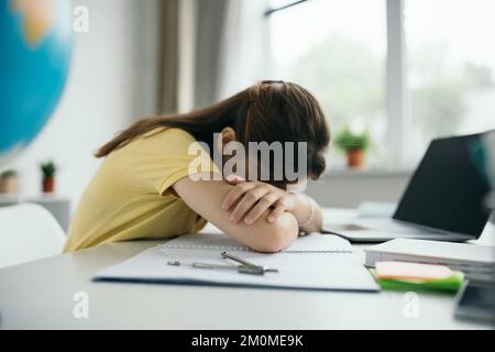 vue latérale d'une fille épuisée dormant sur un copybook près d'un ordinateur portable avec écran vierge sur un arrière-plan flou Banque D'Images