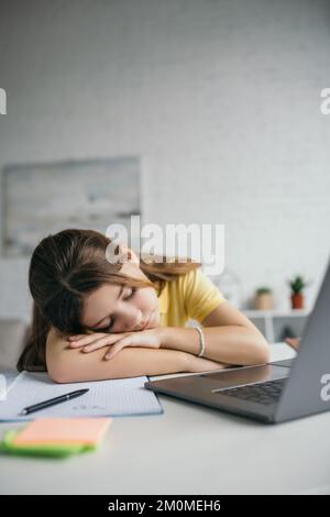 un enfant épuisé dormant près d'un copybook et d'un ordinateur portable sur un premier plan flou à la maison Banque D'Images