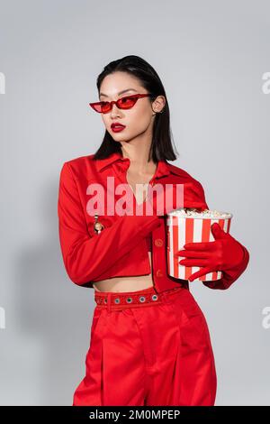 femme asiatique en lunettes de soleil tendance et tenue rouge tenant un seau de pop-corn et regardant loin sur fond gris, image de stock Banque D'Images