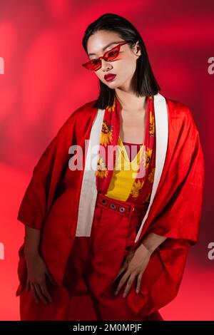 femme asiatique à la mode en kimono cape et lunettes de soleil posant sur fond rouge avec ombre, image de stock Banque D'Images