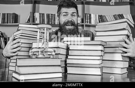 Homme sur le visage heureux entre des piles de livres, tout en étudiant dans la bibliothèque, les étagères sur le fond. Concept de bibliothécaire. Un professeur ou un étudiant avec une barbe est assis Banque D'Images