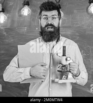 Le professeur de lunettes tient un livre et un microscope. Concept de la science. Un scientifique tient un livre et un microscope, un tableau noir sur fond. Homme avec barbe et Banque D'Images