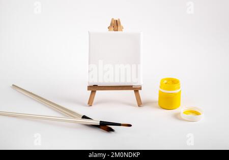 Toile propre, maquette vierge pour la peinture, dessin sur chevalet de trépied de bois, pinceau et gouache jaune dans un pot ouvert. Photo de haute qualité Banque D'Images