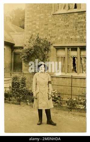 Carte postale originale de WW1 ans d'une jolie fille de terre debout à l'extérieur d'une maison de campagne portant un uniforme de culotte, chapeau de feutre, une tunique de longueur genou, (avec une ceinture intégrée à fermeture à bouton) bras, leggings en cuir et bottes, vers 1917, 1918 R.-U. Banque D'Images