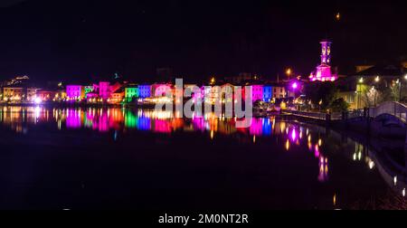 Porto Ceresio, Italie: 12-25-2021: Maisons illuminées par des projecteurs le soir de Noël, par des lumières colorées se reflétant sur l'eau du lac de Luga Banque D'Images