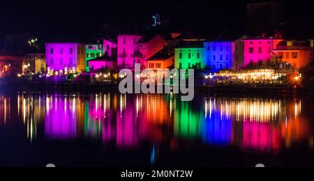 Porto Ceresio, Italie: 12-25-2021: Maisons illuminées par des projecteurs le soir de Noël, par des lumières colorées se reflétant sur l'eau du lac de Luga Banque D'Images