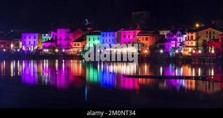 Porto Ceresio, Italie: 12-25-2021: Maisons illuminées par des projecteurs le soir de Noël, par des lumières colorées se reflétant sur l'eau du lac de Luga Banque D'Images