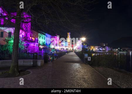 Porto Ceresio, Italie: 12-25-2021: Maisons illuminées par des projecteurs le soir de Noël, par des lumières colorées se reflétant sur l'eau du lac de Luga Banque D'Images