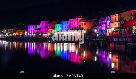 Porto Ceresio, Italie: 12-25-2021: Maisons illuminées par des projecteurs le soir de Noël, par des lumières colorées se reflétant sur l'eau du lac de Luga Banque D'Images