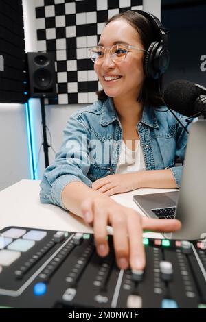Magnifique radio jeune femme très heureuse utilisant un casque et un mélangeur audio tout en diffusant en studio Banque D'Images