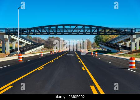 Des bandes récemment peintes et des tonneaux de circulation orange sont visibles sur une route fraîchement recouverte de noir, sous un pont piétonnier. Banque D'Images