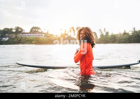 Portrait d'un adolescent noir à poils longs avec planche de surf prête pour le surf avec rétroéclairage au coucher du soleil. Il marche dans les vagues de l'océan Indien. Sports nautiques extrêmes Banque D'Images