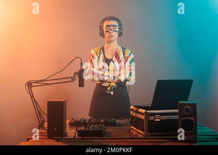 Une femme se posant avec les bras croisés sur les platines, utilisant un casque et un mélangeur pour produire de la musique ou des sons techno. Performances avec le mélangeur et les boutons de commande, l'équipement audio stéréo. Banque D'Images