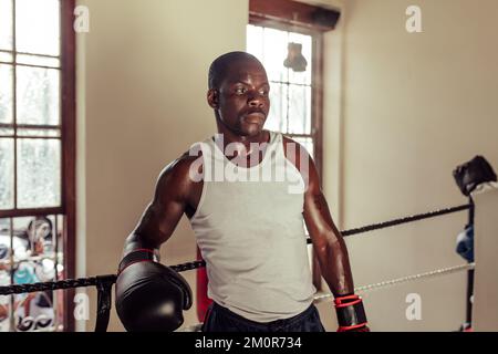 Transpirant jeune homme s'appuyant sur des cordes de boxe en train de prendre une pause de l'entraînement. Un jeune boxeur épuisé dans une salle de boxe. Banque D'Images