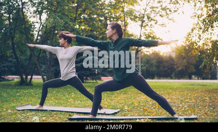 De belles jeunes femmes sont debout dans la posture de la demi-lune Ardha Chandrasana pendant la paire de cours de yoga dans le parc en automne. Mode de vie sain, enseignement et apprentissage et concept sportif. Banque D'Images