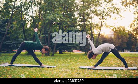 Une étudiante de yoga fait de l'exercice à l'extérieur avec un instructeur qui étire les jambes et se penche vers l'avant, debout sur des tapis de prairie verte et jaune dans le parc de la ville. Banque D'Images