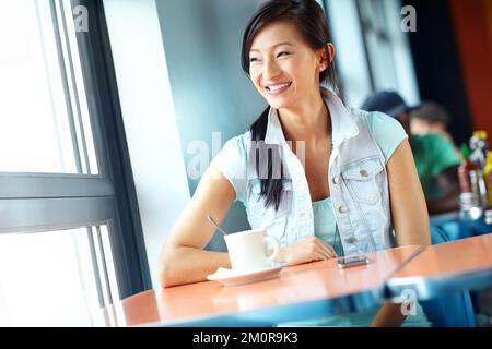 La détente est très agréable. Une belle femme asiatique qui regarde par la fenêtre avec soin tout en appréciant une tasse de café. Banque D'Images