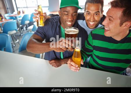 Salut les garçons, buvez. Trois gars amis qui se trinquent leurs bouteilles de bière et leurs verres au bar. Banque D'Images