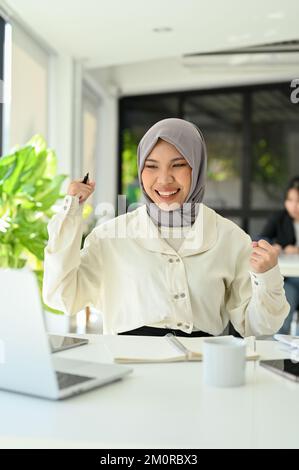 Portrait, une jeune femme de bureau asiatique musulmane se lève, célébrant son succès, recevant de bonnes nouvelles, projet approuvé, obtenir une promotion d'emploi Banque D'Images