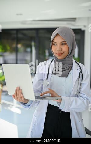 Portrait, belle et professionnelle jeune asiatique musulmane femme médecin en uniforme et hijab à l'aide d'un portable tout en se tenant à l'hôpital. Banque D'Images