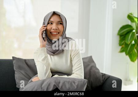 Attrayante et gaie, la jeune femme musulmane asiatique aime parler au téléphone avec son ami tout en se relaxant dans son salon. Banque D'Images