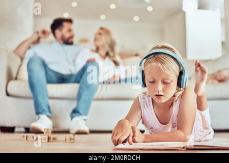 Petite fille caucasienne lisant un livre d'histoire tout en portant des écouteurs à l'écoute de la musique avec ses parents en arrière-plan. Enfant lisant un conte de fées Banque D'Images