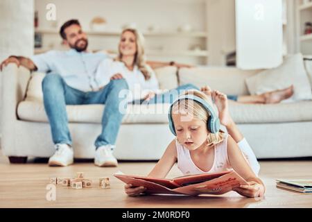 Petite fille caucasienne lisant un livre d'histoire tout en portant des écouteurs à l'écoute de la musique avec ses parents en arrière-plan. Enfant lisant un conte de fées Banque D'Images