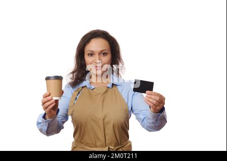 Une serveuse charmante, en tablier de chef, tient une tasse de papier jetable de boisson chaude à emporter et une carte de crédit noire vierge Banque D'Images