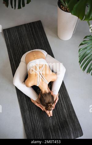 Une jeune fille menant un mode de vie sain et pratiquant le yoga s'entraîne tout en s'asseyant sur un tapis dans des vêtements de sport blancs dans la chambre Banque D'Images