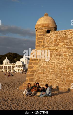 Espagne, Andalousie, Cadix, Caleta Beach, jeune guitariste avec ses amis assis sur le sable face à la mer au pied d'un fort avec l'ancien centre thermal dans le fond dans la lumière chaude de la fin de la journée Banque D'Images