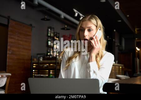 Image d'une femme blonde émotionnellement posant assise à l'intérieur à la caffe en utilisant un ordinateur portable pour le travail. Faites preuve d'une grande surprise en répondant à la question du client. Banque D'Images