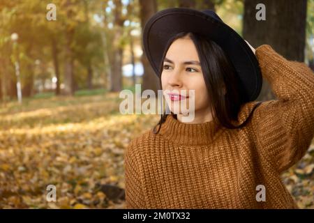 portrait d'une femme élégante et souriante avec de longs cheveux marchant dans le parc vêtu d'un manteau chaud brun automne mode tendance, style de rue portant un chapeau Banque D'Images