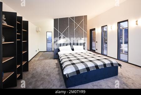 Intérieur minimaliste de la chambre avec grand lit et étagères en bois brun. Vue avant du grand lit, avec couverture en tissu écossais contre le mur gris et décoration géométrique dans la chambre. Concept d'intérieur. Banque D'Images