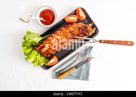 côtes de porc grillées sur une plaque rectangulaire en céramique avec des tranches de tomate fraîches et de la laitue. sauce pour améliorer le goût. vue de dessus. arrière-plan blanc Banque D'Images