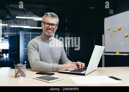 Portrait d'un homme d'affaires senior prospère, d'un homme gris aux cheveux en lunettes souriant et regardant l'appareil photo, d'un patron d'investisseur mature travaillant dans un immeuble de bureau moderne utilisant un ordinateur portable au travail. Banque D'Images