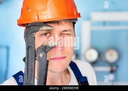 Le plombier professionnel dans le casque regarde directement dans la caméra à travers la grande clé réglable. Grand portrait d'un vrai travailleur de 30 ans.. Banque D'Images