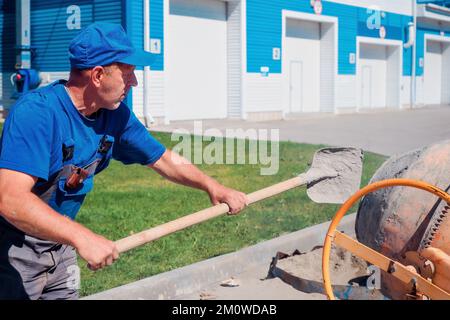 Le travailleur de la construction en uniforme travaille sur le chantier de construction le jour de l'été. La couche de brique de vieille pelle le ciment et le sable dans le mélangeur de béton. Flux de travail authentique... Banque D'Images