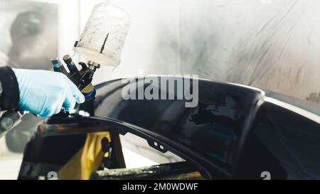 Gros plan en intérieur du processus de vernir les ailes avec peinture automobile noire. Main dans le gant en caoutchouc bleu tenant le pistolet de pulvérisation de peinture. Photo de haute qualité Banque D'Images