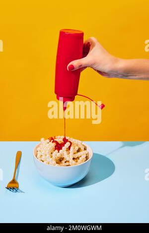 Femelle à la main versant du ketchup dans un bol avec des colliers symbolisant les pâtes, des nouilles sur la nappe bleue sur fond jaune vif. Cuisine pop art Banque D'Images