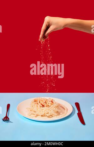 Assiette à saler pour les mains de femmes avec colliers en perles symbolisant les nouilles, pâtes sur nappe bleue sur fond rouge. Photographie d'art pop. Banque D'Images